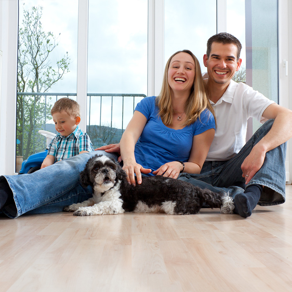 Eine junge Familie mit Hund sitzt auf dem Boden und lacht in die Kamera