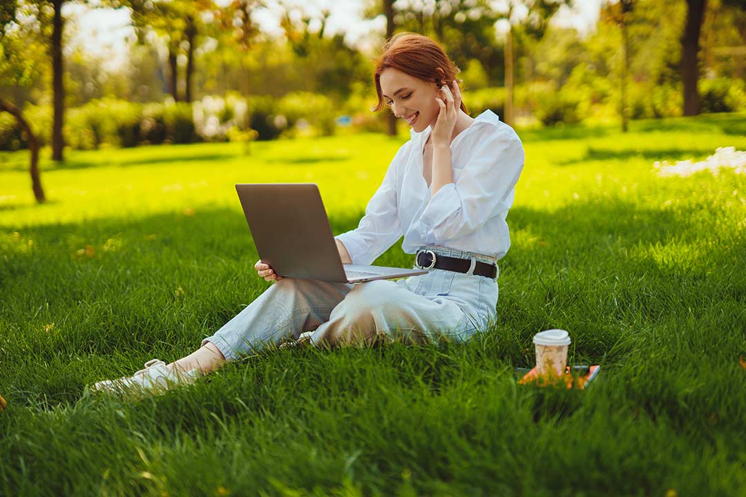 Eine junge rothaarige Frau sitzt in einem Park und arbeitet an einem Laptop